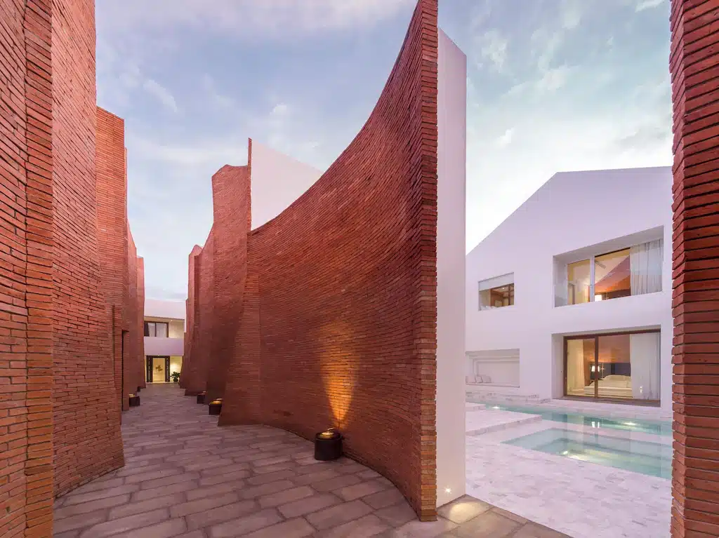 กำแพงอิฐแดงโค้งขนาบทางเดินหิน นำไปสู่ตึกสีขาวทันสมัยที่มีหน้าต่างบานใหญ่และสระว่ายน้ำ—บรรยากาศที่เชิญชวนซึ่งโดดเด่นท่ามกลางสถานที่ท่องเที่ยวในอยุธยา.