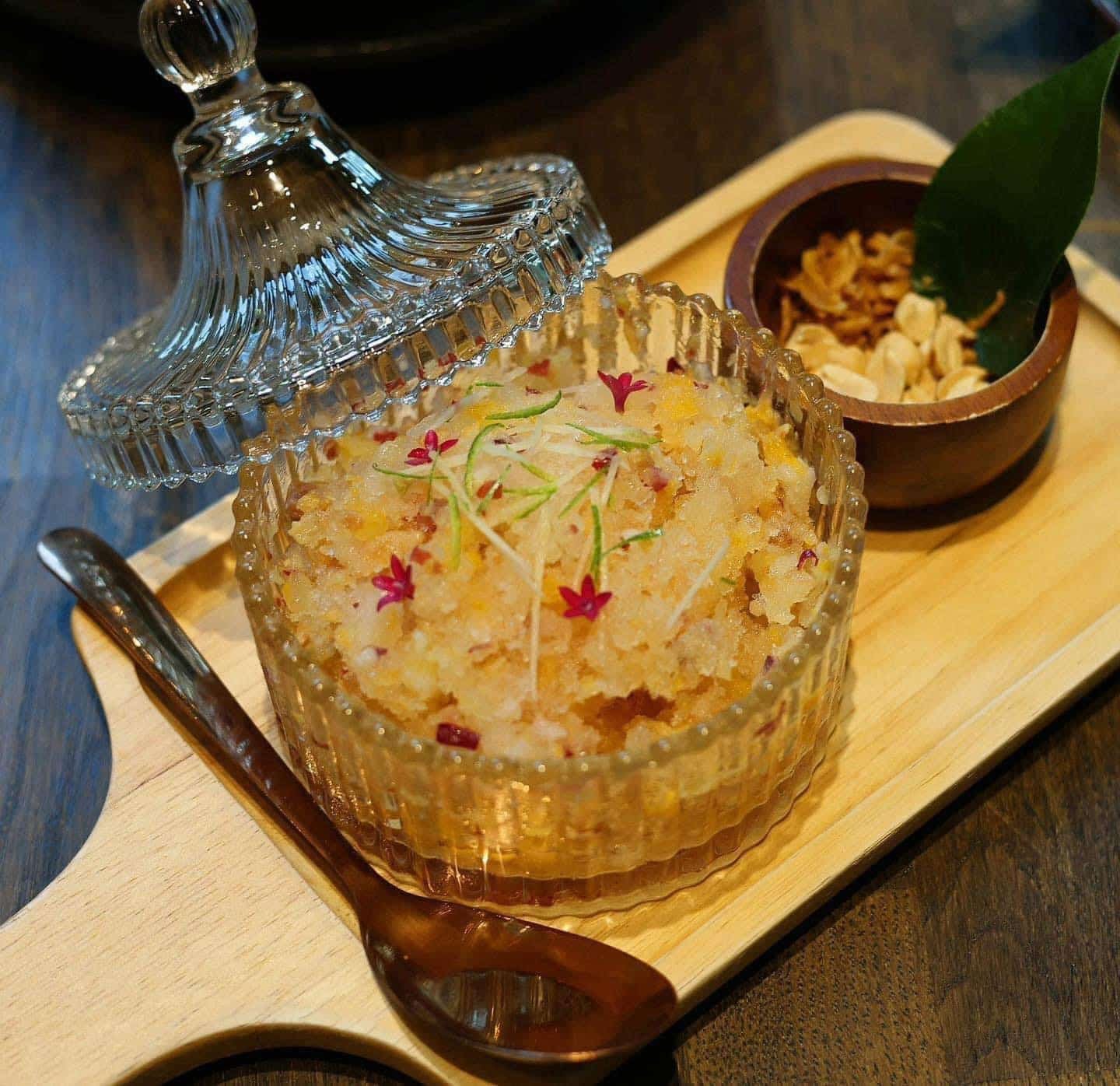 จานแก้วที่มีฝาปิดบรรจุขนมหวานเนื้อละเอียดตกแต่งด้วยสมุนไพรและดอกไม้เล็ก ๆ เสิร์ฟบนถาดไม้พร้อมถั่วและหอมเจียวกรอบ—รสชาติอันน่ารื่นรมย์ที่คุณอาจพบได้เมื่อไปเที่ยวอยุธยา.