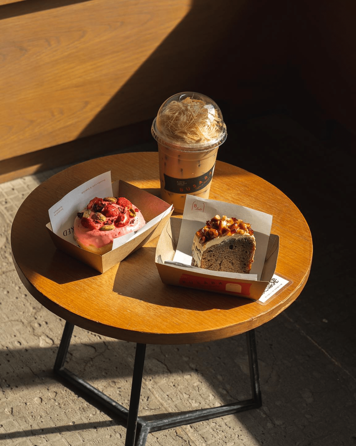 โต๊ะกลมไม้พร้อมกาแฟเย็น หนึ่งชิ้นของขนมอบสีชมพูเคลือบน้ำตาลกับเบอร์รี่ และเค้กหนึ่งชิ้นในแสงแดด—เหมาะสำหรับการผ่อนคลายที่ ที่เที่ยวอยุธยา หรือที่ท่องเที่ยวในอยุธยาที่แสนสบาย.