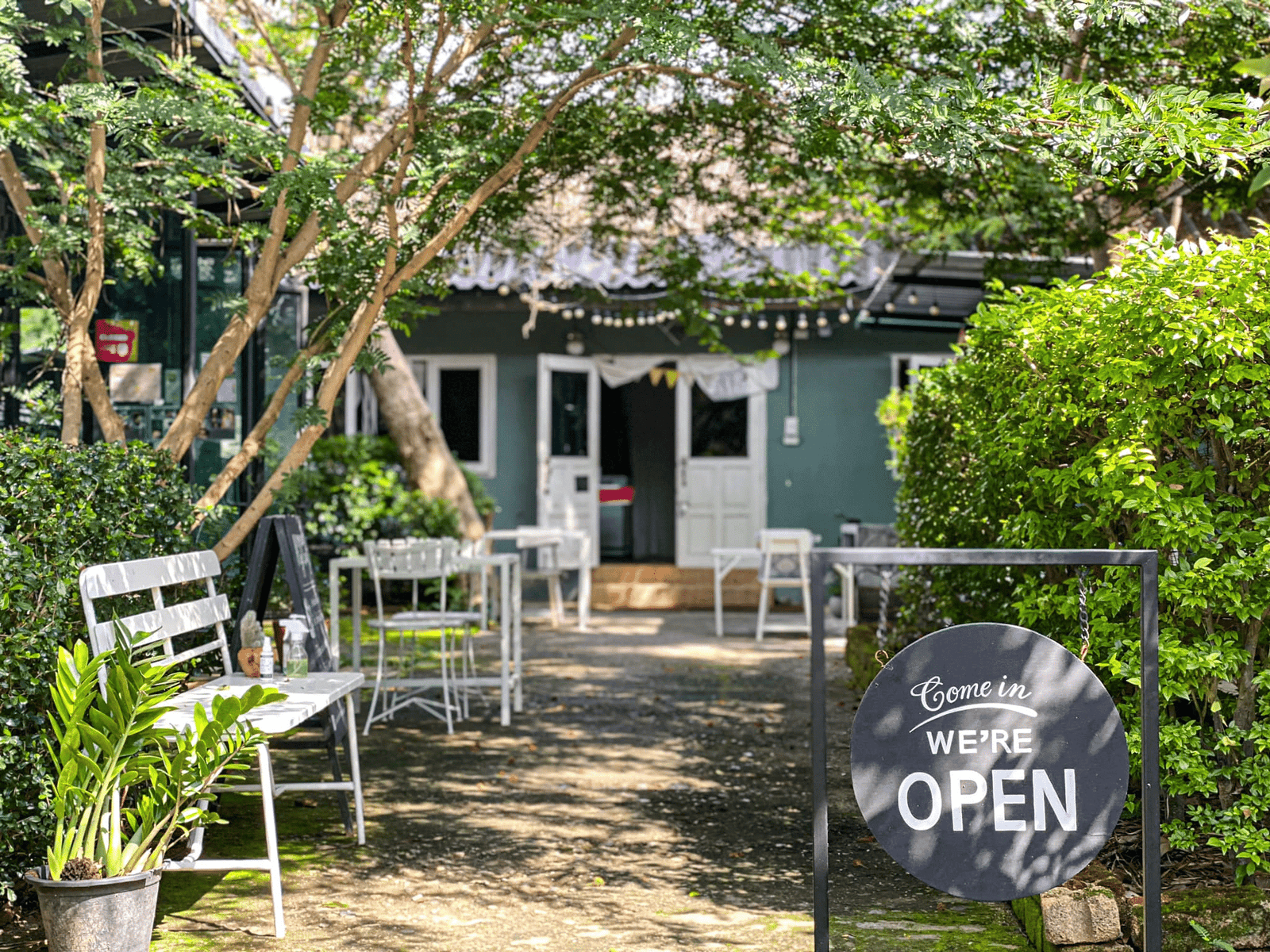 พื้นที่นั่งกลางแจ้งของคาเฟ่ที่มีโต๊ะและเก้าอี้สีขาว, ต้นไม้สีเขียว, และป้ายที่เขียนว่า "เข้ามาเลย, เราเปิดแล้ว" ที่ทางเข้า—เหมาะสำหรับการผ่อนคลายขณะสำรวจ ที่เที่ยวอยุธยา.