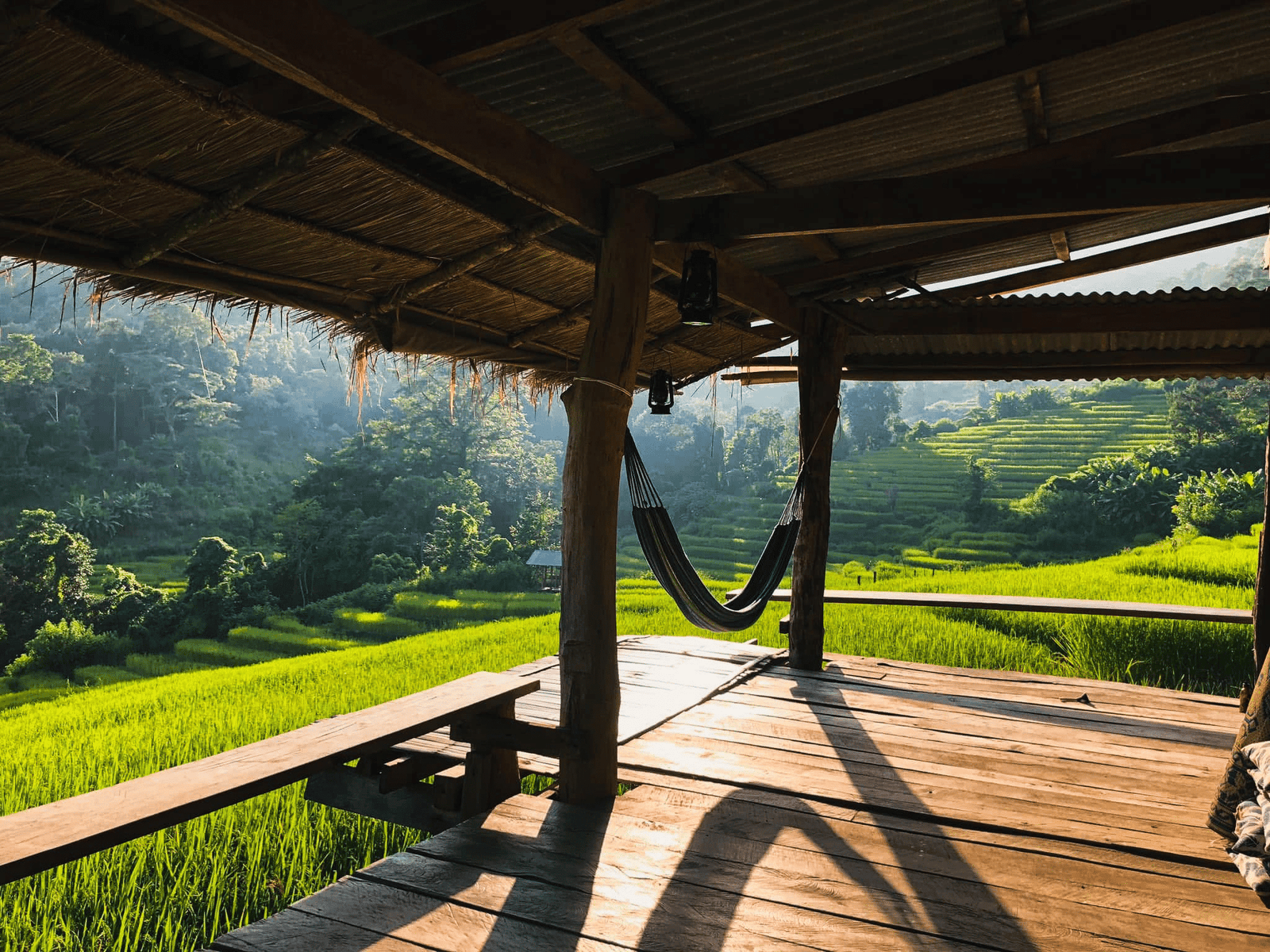 ระเบียงไม้ที่มีเปลญวนแขวนอยู่ มองเห็นทุ่งนาขั้นบันไดสีเขียวและภูเขาที่ปกคลุมด้วยป่าไม้ในระยะไกล ภายใต้แสงแดดอ่อนๆ—สร้างบรรยากาศอันเงียบสงบที่พบได้ในสถานที่ท่องเที่ยวหลายแห่งในอยุธยา.