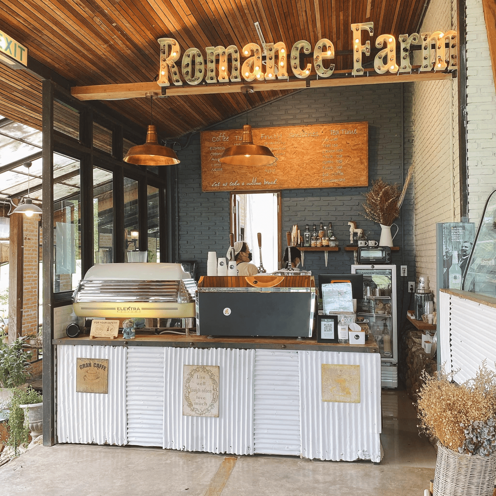 เคาน์เตอร์คาเฟ่ขนาดเล็กที่ Romance Farm สถานที่ท่องเที่ยวที่น่าหลงใหลในอยุธยา มีเครื่องชงกาแฟ ตู้โชว์ เมนูบอร์ด โคมไฟแขวน และการตกแต่งแบบชนบท พร้อมป้ายขนาดใหญ่แขวนอยู่ด้านบน.