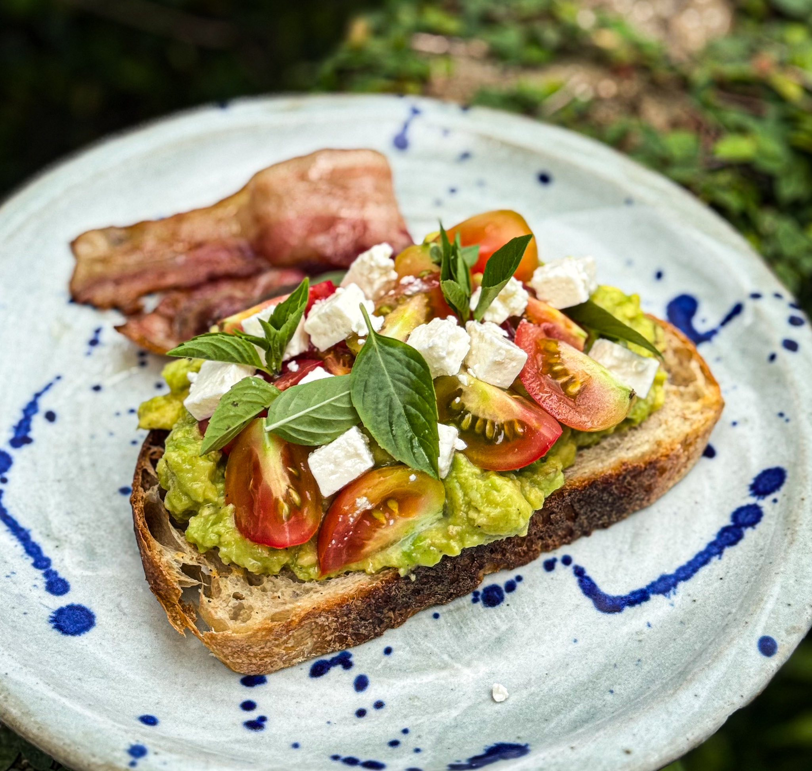 ขนมปังแผ่นหนึ่งทาด้วยอะโวคาโดบด มะเขือเทศเชอร์รี่ ชีสเฟต้า และใบโหระพาสด เสิร์ฟพร้อมเบคอนบนจานสไตล์สวย—เหมาะสำหรับเพลิดเพลินก่อนออกสำรวจสถานที่ท่องเที่ยวอยุธยา หรือผ่อนคลายหลังจากเยี่ยมชมสถานที่ท่องเที่ยวอยุธยา.