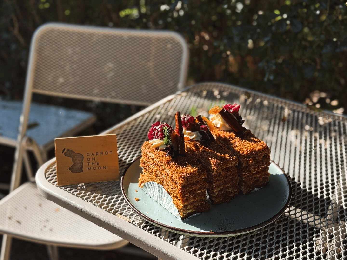 เค้กชั้นสามชิ้นพร้อมผลไม้บนจาน วางบนโต๊ะโลหะกลางแจ้งข้างนามบัตร "แครอทบนดวงจันทร์"—เหมาะสำหรับการผ่อนคลายขณะสำรวจสถานที่ท่องเที่ยวอยุธยา.
