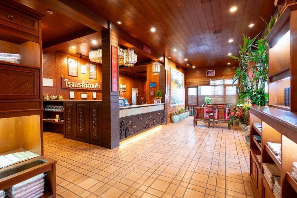 ล็อบบี้โรงแรมที่สว่างไสวด้วยแสงไฟ มีผนังและเพดานไม้ โต๊ะต้อนรับ ชั้นวางผ้าเช็ดตัว ต้นไม้ในกระถาง และพื้นที่นั่งเล่นริมหน้าต่างบานใหญ่—เหมาะสำหรับการพักผ่อนที่ ที่พักน่านในเมือง.
