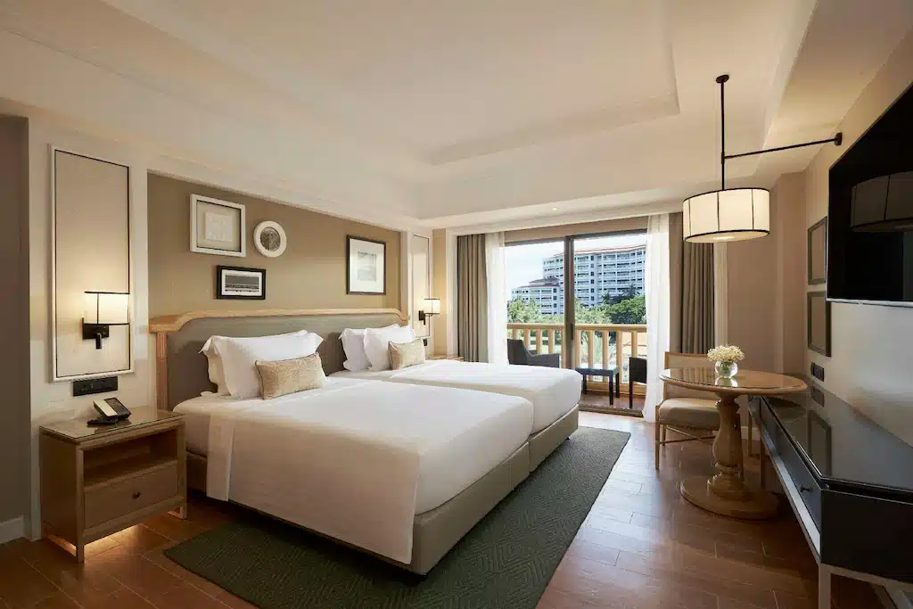 ห้องพักโรงแรมสมัยใหม่พร้อมเตียงคู่สองเตียง โต๊ะข้างเตียง ทีวี และโต๊ะกลมเล็กพร้อมเก้าอี้ใกล้ระเบียงที่มองเห็นวิวสีเขียว—ตัวเลือกที่ยอดเยี่ยมสำหรับ ที่พักหัวหินราคาถูก หรือใครก็ตามที่กำลังมองหา โรงแรมหัวหินใกล้ชิดธรรมชาติ.