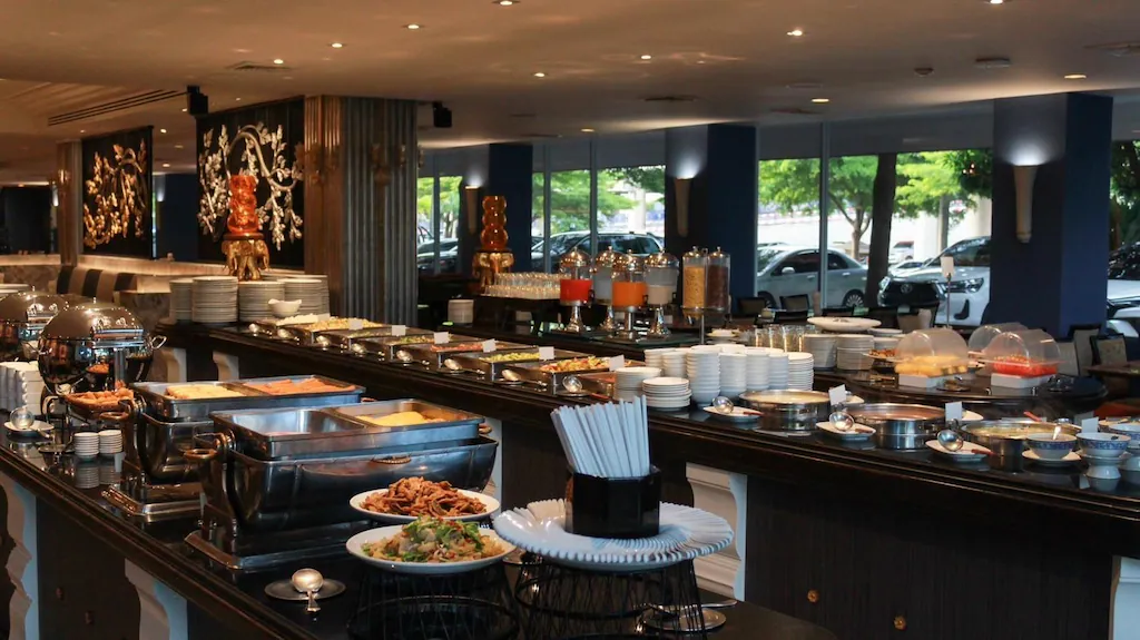 บุฟเฟ่ต์อาหารในร้านอาหารที่มีอาหารหลากหลายชนิด จาน และเครื่องดื่มจัดเรียงอยู่บนเคาน์เตอร์ยาวที่ โรงแรมอยุธยา; หน้าต่างบานใหญ่เผยให้เห็นรถยนต์ที่จอดอยู่และต้นไม้เขียวชอุ่มด้านนอก.