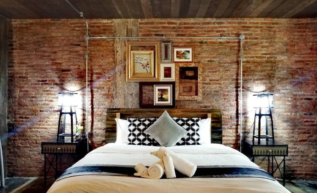 เตียงที่จัดไว้อย่างเรียบร้อยพร้อมหมอนสีขาวดำ ผ้าเช็ดตัวม้วนเป็นรูปทรงสวยงาม และโคมไฟสว่างบนโต๊ะข้างเตียง ตัดกับผนังอิฐเปลือยที่ประดับด้วยกรอบรูป—บรรยากาศอบอุ่นสำหรับการเข้าพักของคุณที่ ที่พักบ่อเกลือ.