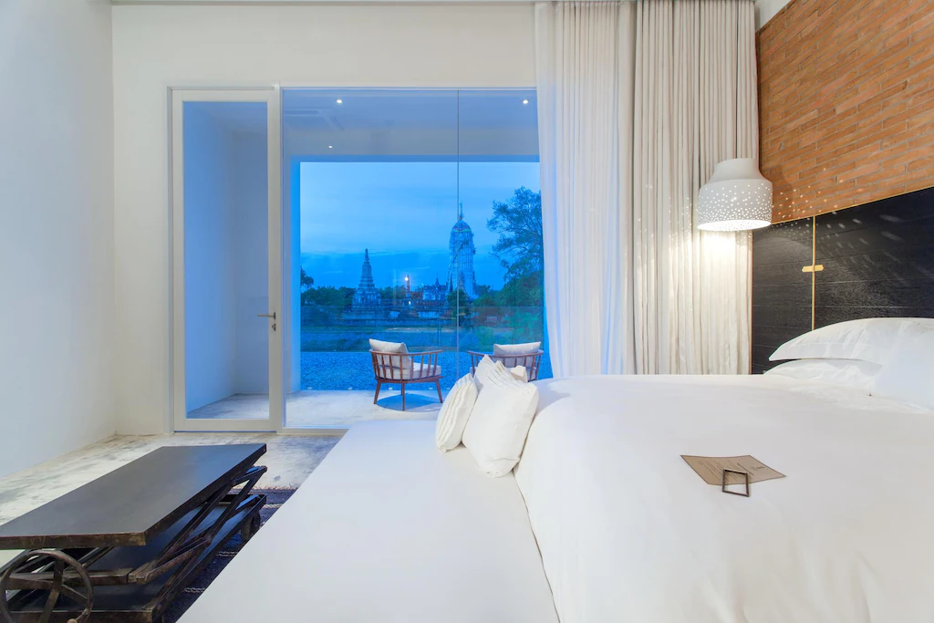 ห้องพักโรงแรมสมัยใหม่พร้อมเตียงที่จัดไว้อย่างเรียบร้อย โต๊ะกาแฟ และเก้าอี้ระเบียงริมหน้าต่างบานใหญ่ที่มองเห็นวัดและต้นไม้ในยามพลบค่ำ—เหมาะสำหรับผู้ที่มองหาที่พักอยุธยาหรือโรงแรมอยุธยาพร้อมวิวที่น่าจดจำ.