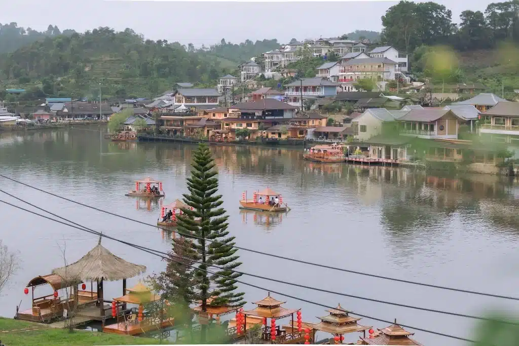 เรือเล็กหลายลำที่มีผู้คนลอยอยู่บนทะเลสาบที่สงบเงียบ รายล้อมไปด้วยบ้านเรือนบนเนินเขาและต้นไม้สีเขียว ภายใต้ท้องฟ้าที่มืดครึ้ม—ทำให้นึกถึงทัศนียภาพที่สงบเงียบที่พบได้ที่ ที่เที่ยวอยุธยา.