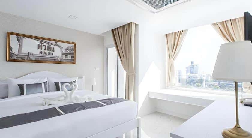 ห้องพักโรงแรมสว่างสดใสพร้อมเตียงขนาดใหญ่ การจัดผ้าขนหนูรูปหงส์ ศิลปะบนผนัง ที่นั่งริมหน้าต่าง และวิวเมืองผ่านหน้าต่างบานใหญ่พร้อมผ้าม่านสีเบจ—เหมาะสำหรับนักเดินทางที่กำลังมองหา ที่พักหัวหินราคาถูก ที่มีความสะดวกสบายและสไตล์.