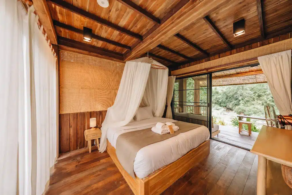 ห้องนอนไม้พร้อมเตียงมีหลังคาคลุม ผ้าปูที่นอนสีขาว ผ้าเช็ดตัววางบนเตียง หน้าต่างบานใหญ่พร้อมผ้าม่าน และวิวต้นไม้สีเขียวด้านนอก—เหมาะสำหรับผู้ที่ต้องการ ที่พักน่านในเมือง หรือ ที่พักปัว ในบรรยากาศเงียบสงบ.