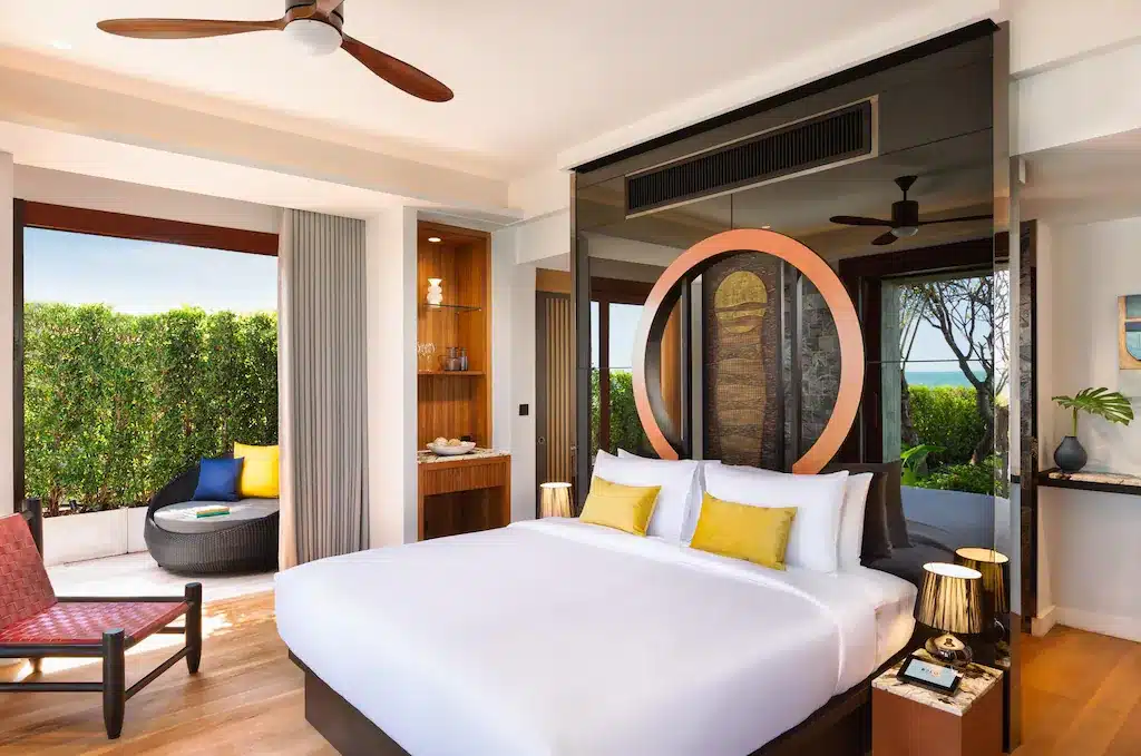 ห้องพักโรงแรมสมัยใหม่พร้อมเตียงขนาดใหญ่ หมอนสีเหลือง พัดลมเพดาน และระเบียงกลางแจ้งส่วนตัวพร้อมที่นั่ง ล้อมรอบด้วยพื้นที่สีเขียว—เหมาะสำหรับการพักผ่อนที่ โรงแรมหัวหิน หรือผู้ที่มองหา ที่พักหัวหินติดทะเล.