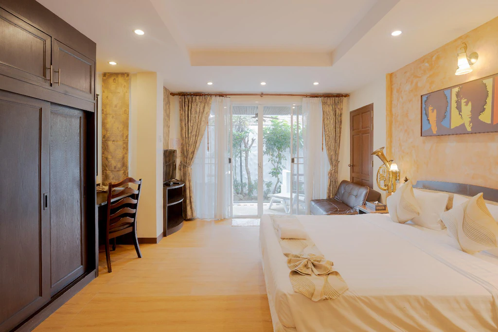 ห้องพักในโรงแรมหัวหินที่มีแสงสว่างเพียงพอ พร้อมเตียงขนาดใหญ่ โซฟา โต๊ะทำงาน ตู้เสื้อผ้า และประตูกระจกที่เปิดออกสู่ระเบียงซึ่งมีต้นไม้สีเขียวอยู่ด้านนอก.