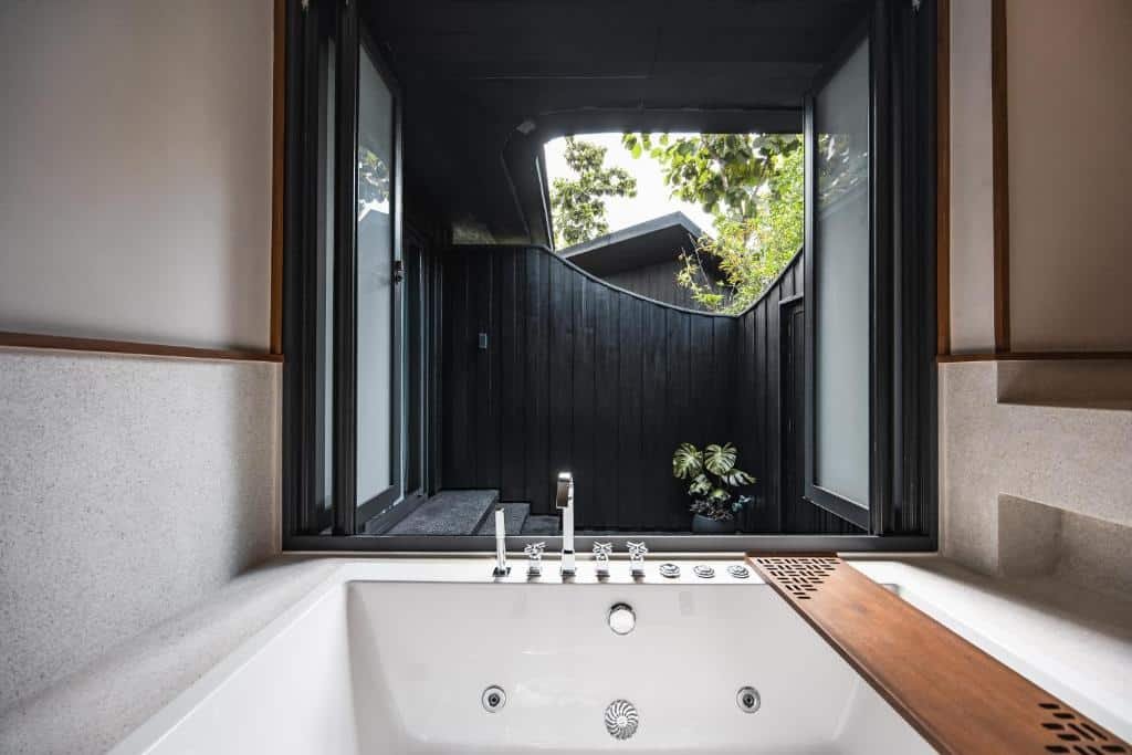 อ่างอาบน้ำสมัยใหม่พร้อมระบบน้ำวนตั้งอยู่ใต้หน้าต่างบานเปิด ให้ทัศนียภาพของพื้นที่กลางแจ้งที่มีผนังไม้สีเข้มและพืชพรรณสีเขียว—เหมาะอย่างยิ่งสำหรับการผ่อนคลายที่ ที่พักบ่อเกลือ อันเงียบสงบ.