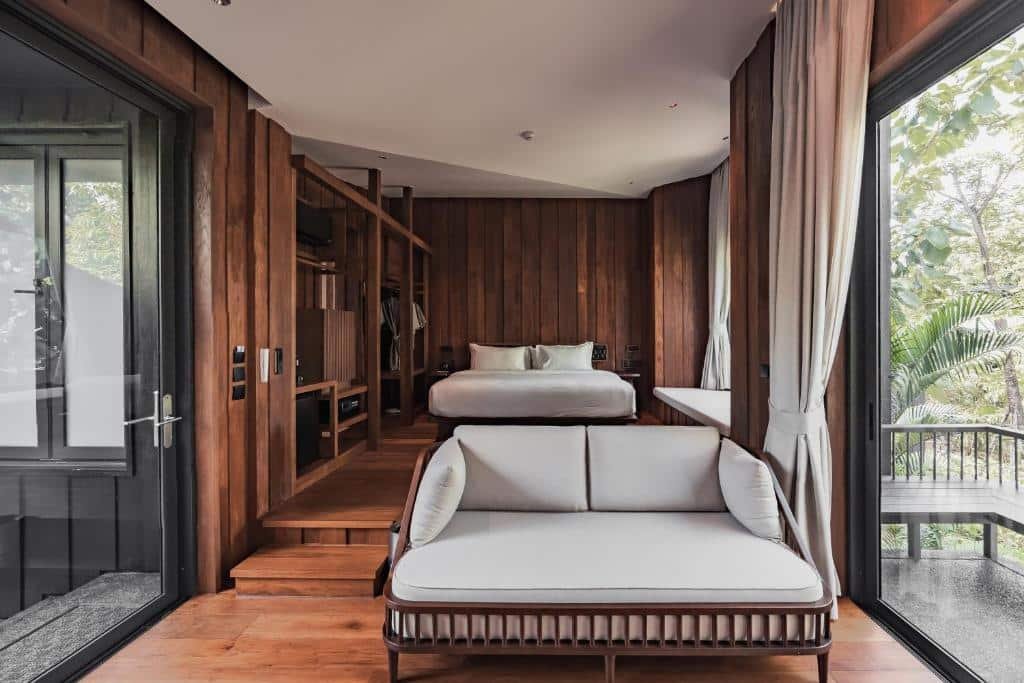 ห้องนอนสไตล์โมเดิร์นพร้อมผนังไม้, เตียงสีขาว, ม้านั่งบุฟองน้ำ, หน้าต่างบานใหญ่, และแสงธรรมชาติที่มองเห็นต้นไม้เขียวขจี—เหมาะสำหรับผู้ที่ต้องการ ที่พักน่าน หรือ ที่พักน่านในเมือง ที่เงียบสงบ.