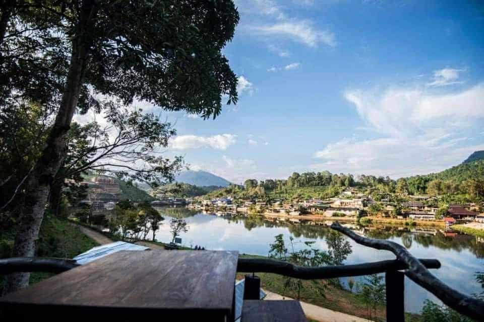 โต๊ะไม้ตั้งอยู่เหนือแม่น้ำที่สงบเงียบ มีเงาสะท้อนของต้นไม้และบ้านเรือนรายล้อมไปด้วยความเขียวขจีและภูเขาภายใต้ท้องฟ้าที่มีเมฆบางส่วน—เป็นภาพที่ชวนให้ใครก็ตามที่ต้องการเที่ยวอยุธยาและค้นพบสถานที่ท่องเที่ยวในอยุธยา.