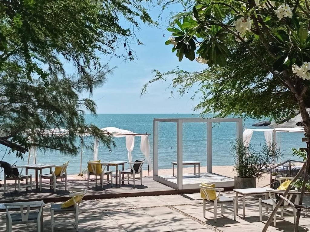 พื้นที่นั่งเล่นริมชายหาดกลางแจ้งพร้อมโต๊ะและเก้าอี้สีขาว ให้ร่มเงาจากต้นไม้ หันหน้าสู่ทะเลภายใต้ท้องฟ้าสีครามใส—เหมาะอย่างยิ่งสำหรับการพักผ่อนที่โรงแรมหัวหิน หรือค้นหาที่พักหัวหินราคาประหยัดตามแนวชายฝั่ง.