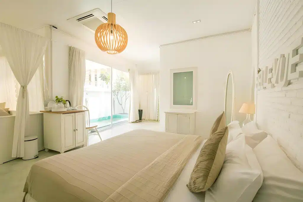ห้องนอนสว่างไสว ทันสมัย พร้อมผ้าปูที่นอนสีเบจ ผนังสีขาว หน้าต่างขนาดใหญ่ โคมไฟแขวน และวิวสระว่ายน้ำภายนอก—เหมาะสำหรับผู้ที่ต้องการที่พักหัวหินราคาถูก ที่มีความสไตล์และความสะดวกสบาย.