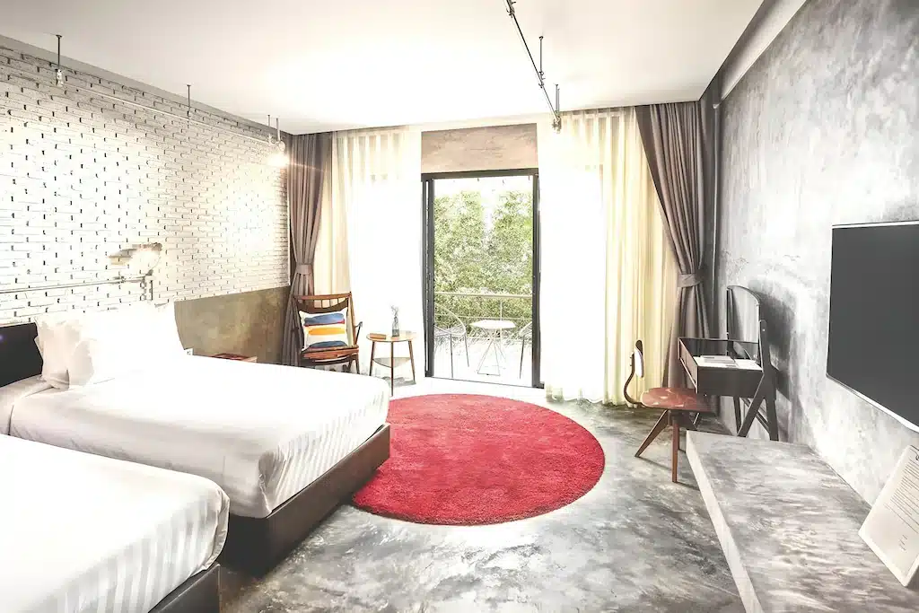 ที่พักหัวหินราคาประหยัดสไตล์โมเดิร์น ประกอบด้วยเตียงสองเตียง พรมแดงทรงกลม เฟอร์นิเจอร์มินิมอล โทรทัศน์จอใหญ่ติดผนังคอนกรีต และระเบียงเล็กพร้อมเก้าอี้สำหรับนั่งพักผ่อนรับลมทะเล.