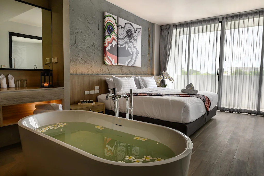 ห้องพักโรงแรมสมัยใหม่ที่ โรงแรมหัวหิน พร้อมอ่างอาบน้ำแบบลอยตัวพร้อมดอกไม้ เตียงขนาดใหญ่ งานศิลปะที่มีสไตล์ และหน้าต่างบานใหญ่จากพื้นจรดเพดานพร้อมผ้าม่านบางเบา—เหมาะสำหรับผู้ที่มองหา ที่พักหัวหินติดทะเล.