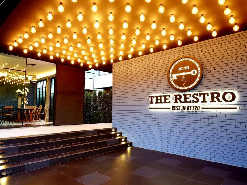 ทางเข้า "The Restro" ร้านอาหารสุดชิคใกล้ที่พักหัวหิน โดดเด่นด้วยผนังอิฐสีขาว ป้ายไฟสว่าง บันได และเพดานประดับด้วยหลอดไฟทรงกลม มองเห็นพื้นที่รับประทานอาหารอันแสนอบอุ่นผ่านหน้าต่างกระจกบานใหญ่.
