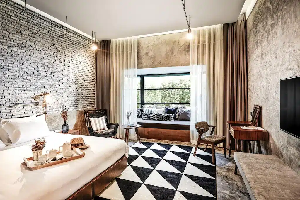 ห้องพักโรงแรมสมัยใหม่พร้อมผนังอิฐเปลือย พรมลายเรขาคณิต เตียงขนาดใหญ่พร้อมถาดอาหารเช้า—เหมาะสำหรับการเข้าพักที่โรงแรมหัวหิน เพลิดเพลินกับที่นั่งริมหน้าต่าง โต๊ะไม้ เก้าอี้สองตัว และทีวีจอแบนในความสะดวกสบายอย่างมีสไตล์.