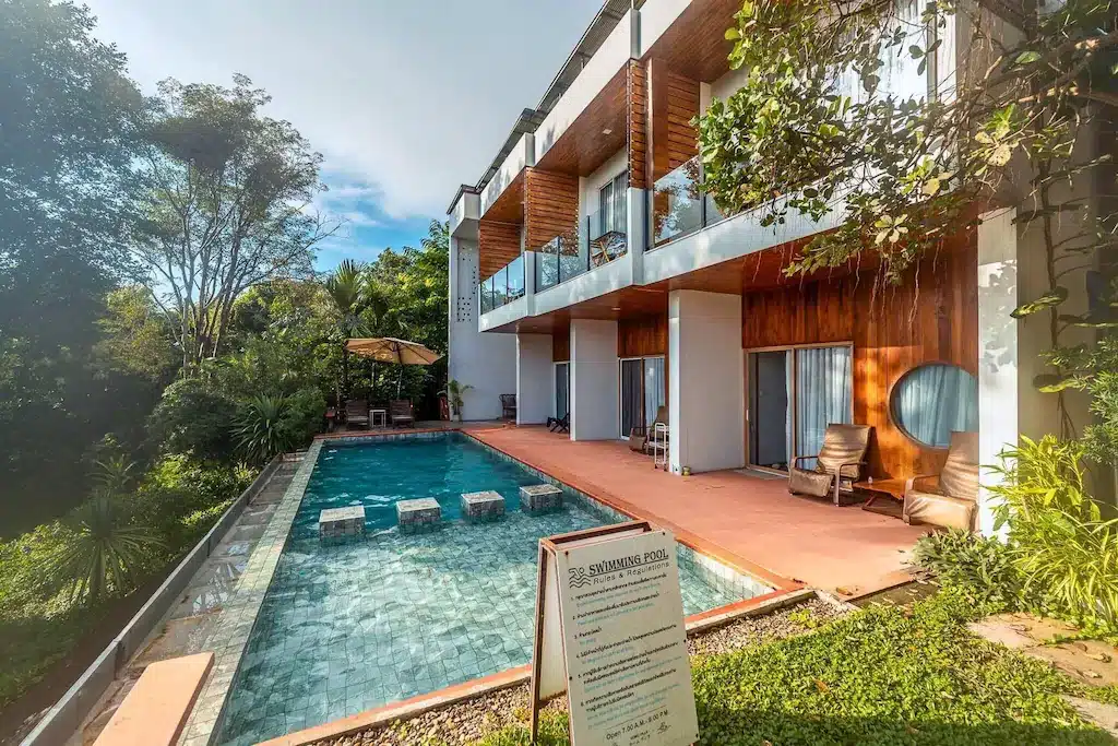 อาคารสองชั้นสมัยใหม่พร้อมระเบียงมองเห็นสระว่ายน้ำกลางแจ้งรูปสี่เหลี่ยมผืนผ้า ล้อมรอบด้วยเก้าอี้ลานและต้นไม้เขียวชอุ่ม เหมาะสำหรับผู้ที่มองหาที่พักในตัวเมืองน่าน พร้อมป้ายข้อมูลอยู่ด้านหน้า.