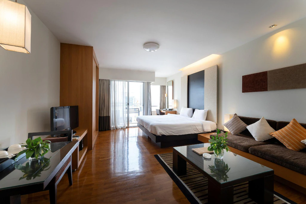 ห้องพักโรงแรมสมัยใหม่พร้อมเตียงขนาดใหญ่ โซฟา โต๊ะกาแฟ โทรทัศน์จอแบน พืช และหน้าต่างบานใหญ่จากพื้นจรดเพดานพร้อมวิวเมือง—เหมาะสำหรับผู้ที่มองหาที่พักในอยุธยาที่มีสไตล์.