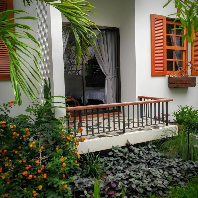 ลานระเบียงขนาดเล็กพร้อมราวเหล็กสีดำ ประตูกระจก และบานหน้าต่างสีส้ม; สวนที่มีต้นไม้สีเขียวและดอกไม้สีส้มอยู่ด้านหน้า—บรรยากาศที่เชิญชวนสำหรับการพักผ่อนที่ ที่พักน่าน.