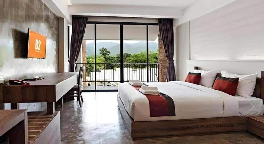 ห้องพักโรงแรมสมัยใหม่พร้อมเตียงคู่ โต๊ะทำงาน โทรทัศน์ติดผนัง และระเบียงที่มองเห็นวิวต้นไม้และภูเขา—เหมาะสำหรับการพักผ่อนหลังจากสำรวจสถานที่ท่องเที่ยวในอยุธยาหรือสถานที่ท่องเที่ยวอื่นๆ ในบริเวณใกล้เคียง.