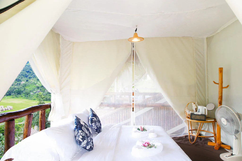 เตียงหลังคาพร้อมผ้าปูที่นอนสีขาวและหมอนลายดอกไม้ตั้งอยู่ในห้องเต็นท์ที่มีม่านบางเบา, ราวแขวนเสื้อไม้, ชุดน้ำชา, และพัดลม เพลิดเพลินกับพืชพรรณเขียวชอุ่มภายนอก—เหมาะสำหรับผู้ที่มองหาที่พักในปัวหรือการพักผ่อนที่เงียบสงบใกล้กับน่าน.