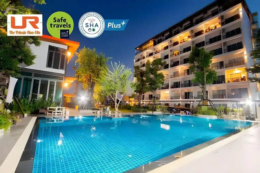 สระว่ายน้ำกลางแจ้งในยามค่ำคืนล้อมรอบด้วยอาคารโรงแรมสมัยใหม่ เหมาะสำหรับการพักผ่อนที่ ที่พักหัวหินติดทะเล พร้อมโลโก้ความปลอดภัยและมาตรฐานแสดงไว้ที่มุมบนซ้าย.