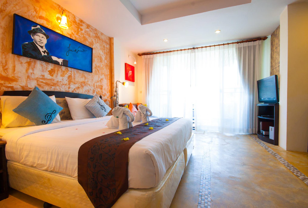 ห้องพักโรงแรมที่แสนสบายที่ โรงแรมหัวหิน พร้อมเตียงคู่ ผ้าเช็ดตัวรูปช้างสองผืน โทรทัศน์ หน้าต่างบานใหญ่พร้อมผ้าม่านบาง และภาพวาดชายในหมวก—เหมาะสำหรับผู้ที่ต้องการ ที่พักหัวหิน ใกล้ทะเล ที่มีสไตล์.