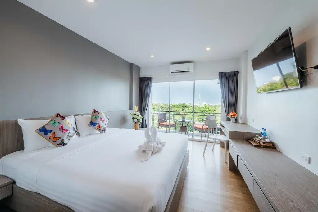 ห้องพักโรงแรมหัวหินสมัยใหม่ พร้อมเตียงที่จัดไว้อย่างเรียบร้อย หมอนผีเสื้อ ศิลปะผ้าขนหนู โต๊ะทำงาน โทรทัศน์จอแบน และระเบียงที่มองเห็นวิวสีเขียว เหมาะสำหรับนักเดินทางที่มองหาความสะดวกสบายในบรรยากาศที่มีเสน่ห์.