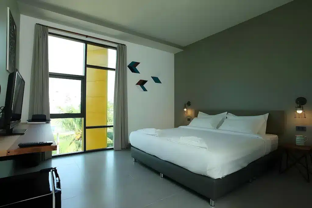 ห้องพักโรงแรมหัวหินสมัยใหม่พร้อมเตียงขนาดใหญ่ ทีวีติดผนัง โต๊ะทำงาน และหน้าต่างสองบานพร้อมผ้าม่านที่เปิดรับแสงธรรมชาติ การตกแต่งเรียบง่ายประกอบด้วยศิลปะบนผนังรูปทรงเรขาคณิตและโคมไฟข้างเตียงเพื่อการพักผ่อนที่ผ่อนคลาย.