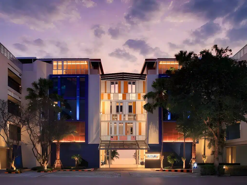 อาคารหลายชั้นสมัยใหม่พร้อมหน้าต่างบานใหญ่และทางเข้าที่เป็นกระจก ล้อมรอบด้วยต้นไม้ ถ่ายในยามพลบค่ำกับท้องฟ้าสีม่วง—โรงแรมหัวหินที่มีสไตล์ เหมาะสำหรับผู้ที่มองหาความสะดวกสบายในบรรยากาศที่สวยงาม.