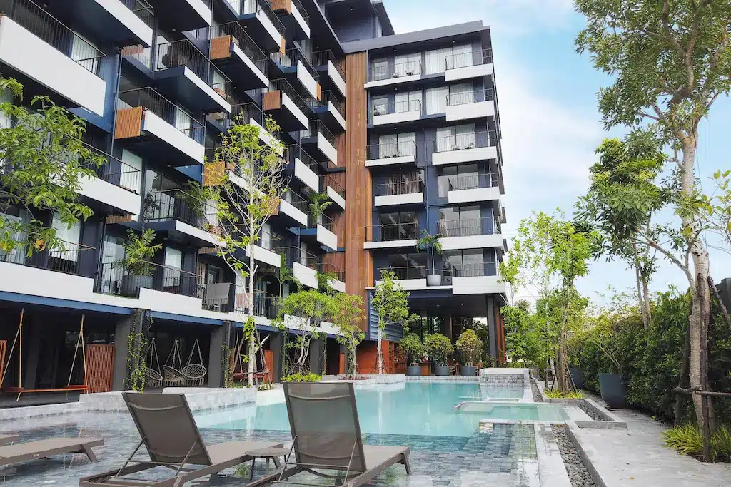 อาคารอพาร์ตเมนต์สมัยใหม่พร้อมระเบียงมองเห็นสระว่ายน้ำที่ล้อมรอบด้วยเก้าอี้เลานจ์และพื้นที่สีเขียว—เหมาะสำหรับผู้ที่มองหาที่พักหัวหินราคาถูกในวันที่แดดสดใส.