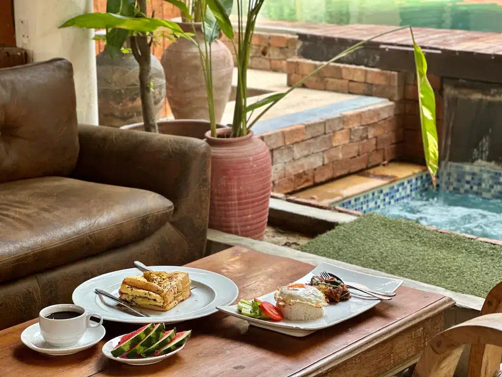 โต๊ะไม้ข้างสระน้ำที่ ที่พักอยุธยาริมน้ำ มีถ้วยกาแฟ น้ำแตงโมหั่นเป็นชิ้น แซนด์วิช ข้าวกับปลา และผักอยู่ บนโซฟาหนังใกล้ ๆ มีต้นไม้ในกระถางสร้างบรรยากาศผ่อนคลาย.