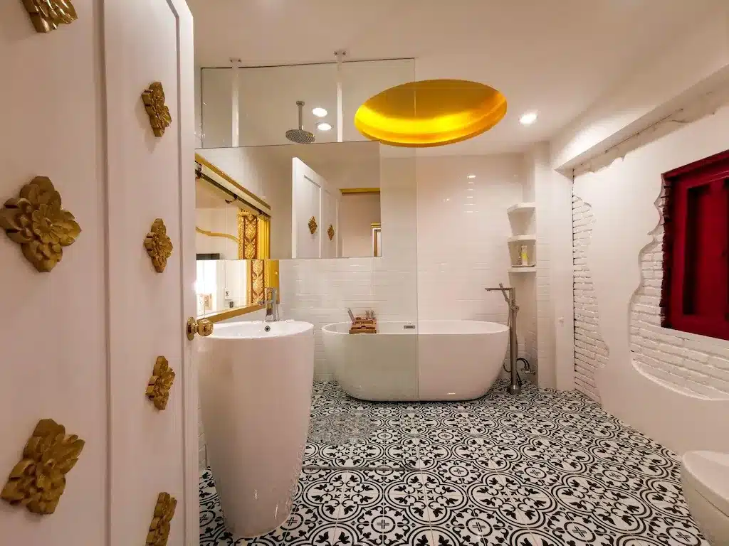 ห้องน้ำที่มีพื้นกระเบื้องลายขาวดำ, อ่างอาบน้ำแบบลอยตัว, อ่างล้างหน้าแบบตั้งบนขา, ตกแต่งด้วยสีทอง, ผนังสีขาว, และหน้าต่างกรอบสีแดง—เหมาะสำหรับโรงแรมอยุธยาที่ต้องการสไตล์ที่อมตะและหรูหราแบบทันสมัย.