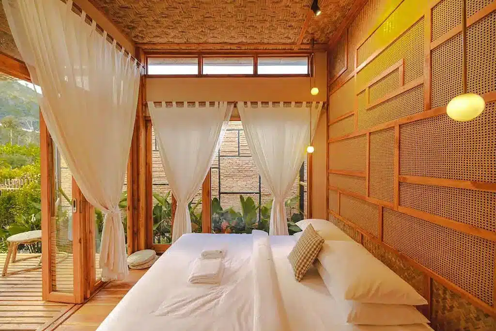 ห้องนอนที่แสนอบอุ่นที่ ที่พักปัว พร้อมเตียงขนาดใหญ่ ผ้าปูที่นอนสีขาว ผ้าม่านบางเบา ผนังไม้ และแสงไฟที่อบอุ่น มองเห็นต้นไม้สีเขียวผ่านประตูและหน้าต่างกระจก.