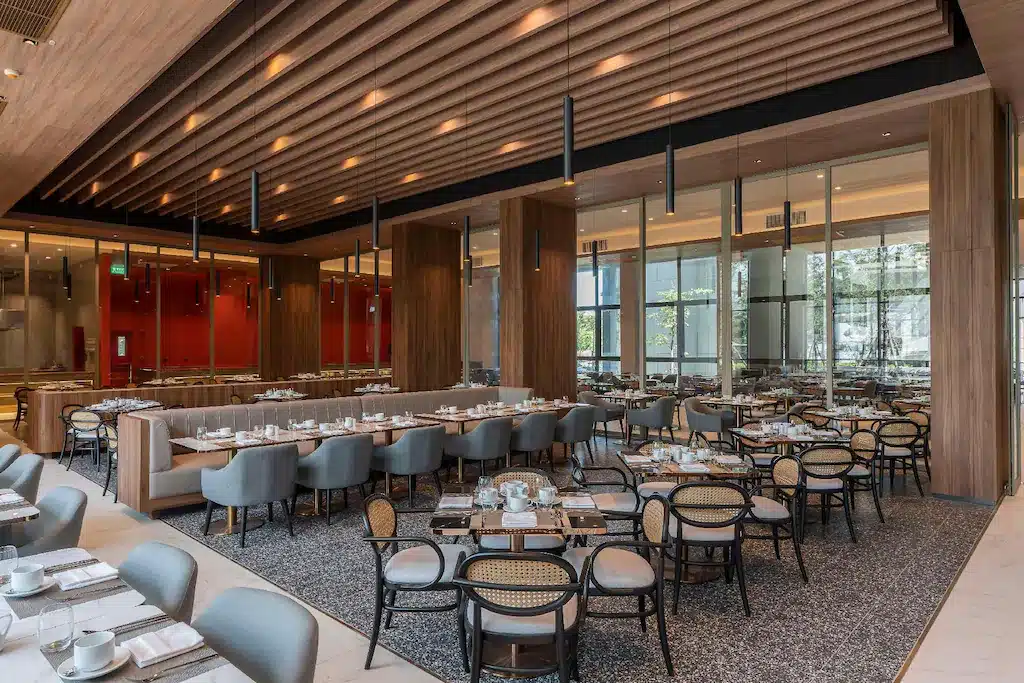 พื้นที่รับประทานอาหารของร้านอาหารสมัยใหม่ที่มีการจัดโต๊ะและเก้าอี้อย่างเป็นระเบียบ เพดานไม้ หน้าต่างบานใหญ่ และแสงไฟที่ผสมผสานระหว่างธรรมชาติและแสงไฟประดิษฐ์—เหมาะอย่างยิ่งสำหรับแขกที่เข้าพักที่ โรงแรมหัวหิน หรือ ที่พักหัวหินติดทะเล.