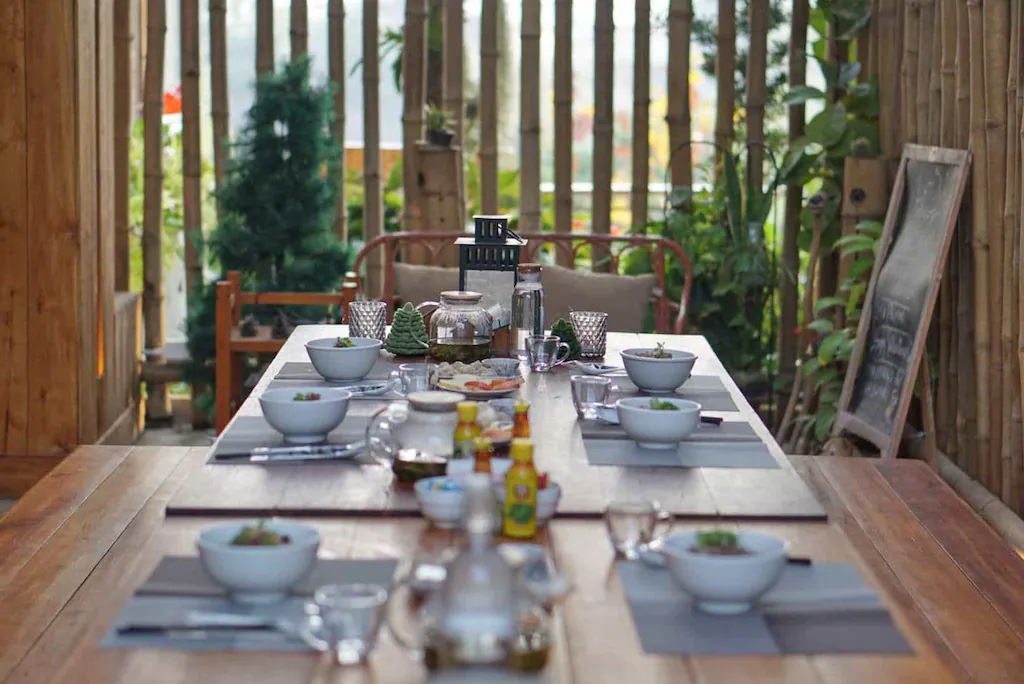 โต๊ะอาหารไม้ถูกจัดเตรียมไว้พร้อมชาม, จาน, แก้ว, และเครื่องปรุงในบริเวณกลางแจ้งที่มีต้นไม้และรั้วไม้ล้อมรอบ—เหมาะสำหรับการรับประทานอาหารที่ ที่พักปัว ท่ามกลางธรรมชาติ.