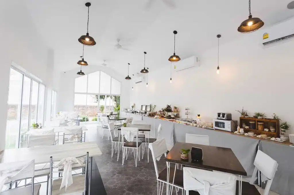 ภายในคาเฟ่ที่สว่างและทันสมัยด้วยผนังสีขาว โต๊ะและเก้าอี้สีขาว การจัดบุฟเฟ่ต์อยู่ทางขวา หน้าต่างบานใหญ่เปิดรับแสงธรรมชาติ—เหมาะสำหรับการพักผ่อนหลังจากสำรวจ ที่พักน่านในเมือง.