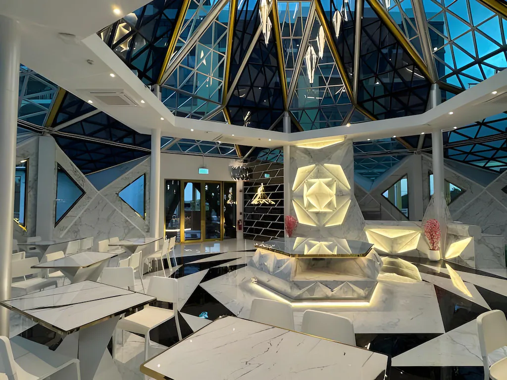 ร้านอาหารสมัยใหม่ภายในตกแต่งด้วยเพดานกระจกทรงเรขาคณิต, การตกแต่งด้วยหินอ่อนมุมเหลี่ยม, โต๊ะและเก้าอี้สีขาว, และผนังที่ประดับด้วยแสงไฟ—เหมาะสำหรับแขกที่กำลังมองหา โรงแรมอยุธยา หรือ ที่พักอยุธยาริมน้ำ ในบรรยากาศที่มีสไตล์.