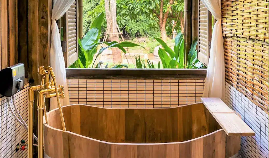 อ่างอาบน้ำไม้พร้อมอุปกรณ์สีทองตั้งอยู่ใต้หน้าต่างเปิดโล่ง มอบวิวที่เงียบสงบของต้นไม้และพืชสีเขียว—เหมาะสำหรับการผ่อนคลายหลังจากสำรวจ ที่พักน่าน ผนังตกแต่งด้วยกระเบื้องสีขาวและไม้ไผ่ล้อมรอบอ่างอาบน้ำ.