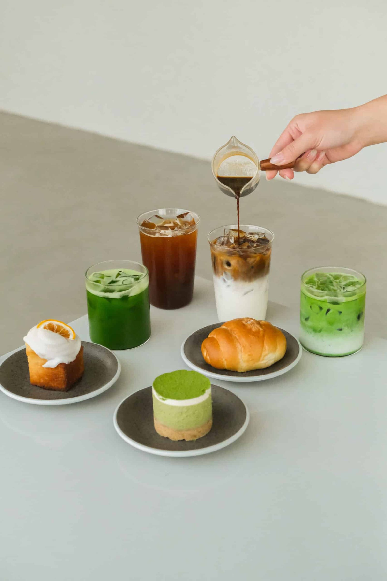 มือกำลังรินเอสเพรสโซ่ลงในแก้วนมบนโต๊ะที่มีเครื่องดื่มเย็น, ขนมอบ, และของหวาน—เหมาะสำหรับการผ่อนคลายก่อนออกไปสำรวจสถานที่ท่องเที่ยวเชียงใหม่หรือวางแผนการผจญภัยครั้งต่อไปของคุณที่เชียงใหม่.