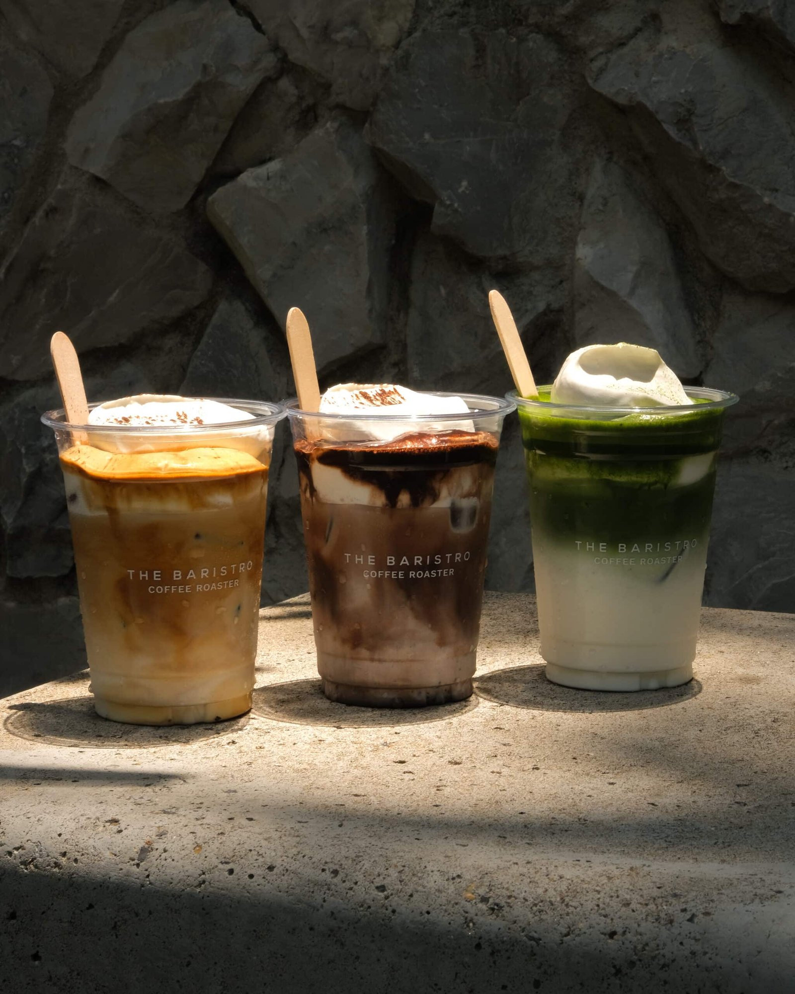 เครื่องดื่มเย็นสามแก้วในถ้วยพลาสติกใสพร้อมวิปครีมและไม้คน วางบนพื้นหินในแสงแดด ติดกับผนังหินที่มีพื้นผิว—เหมาะสำหรับการเพลิดเพลินขณะสำรวจ ที่เที่ยวเชียงใหม่.