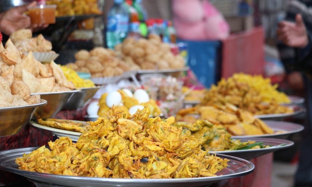 จานของขนมทอดหลากหลายชนิด, ไข่ต้ม, และอาหารริมทางอื่น ๆ ถูกจัดแสดงบนโต๊ะของผู้ขายในตลาดกลางแจ้ง สะท้อนบรรยากาศคึกคักของสถานที่ท่องเที่ยวอยุธยา.