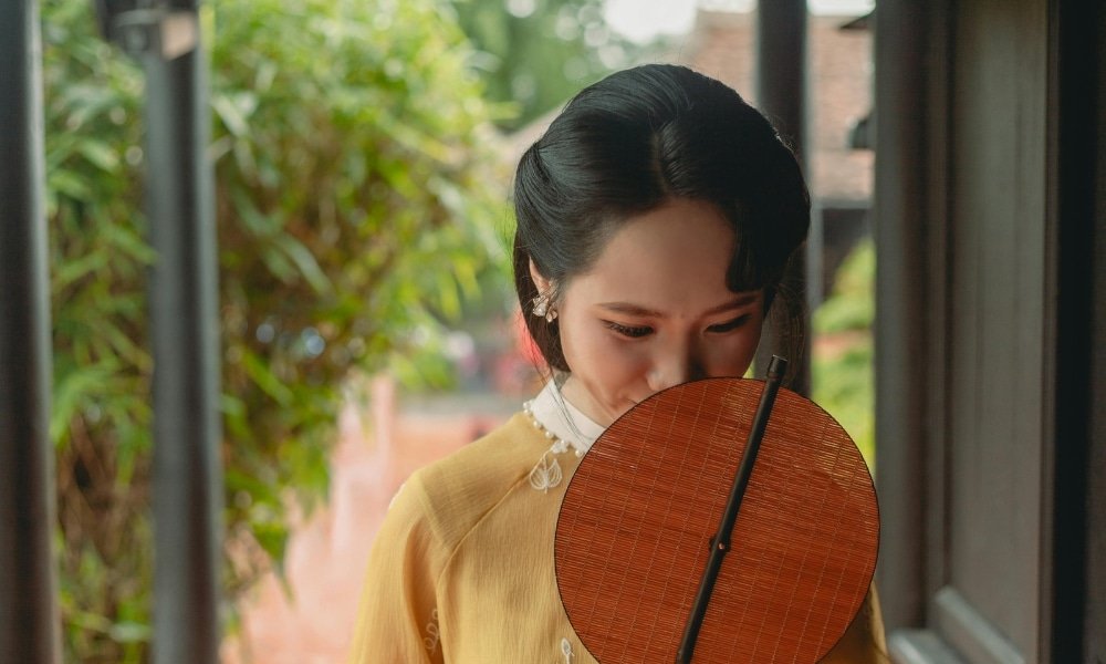 ผู้หญิงในชุดประจำชาติสีเหลืองถือพัดไม้กลมที่ปิดหน้าบางส่วนขณะยืนอยู่ในทางเดินกลางแจ้งที่มีร่มเงา สะท้อนเสน่ห์อันเงียบสงบของสถานที่ท่องเที่ยวอยุธยา.